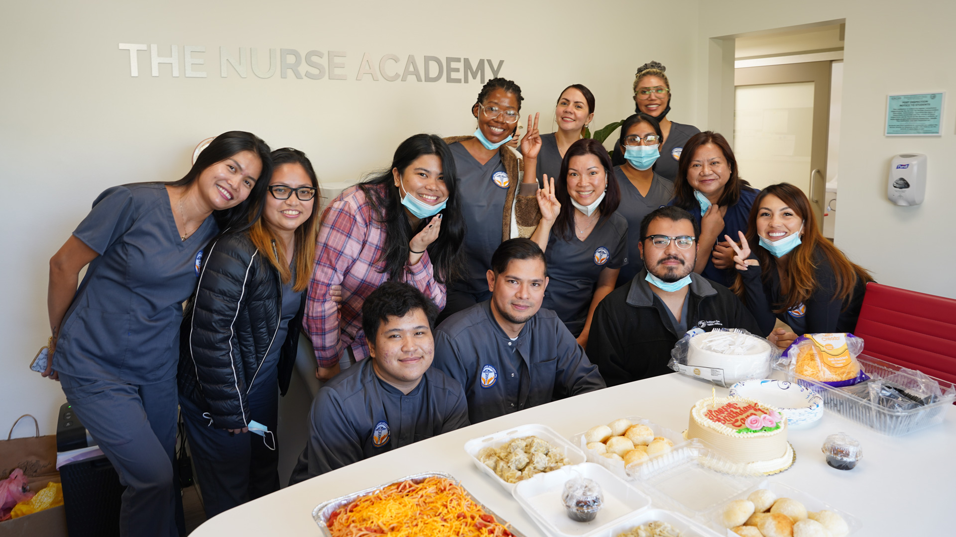 A group of nursing students from The Nurse Academy posing for a photo with a celebratory spread of food on the table.
